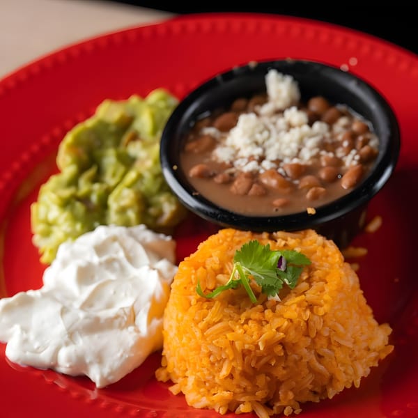A vibrant red plate holds Mexican rice with cilantro, refried beans, guacamole, and sour cream.