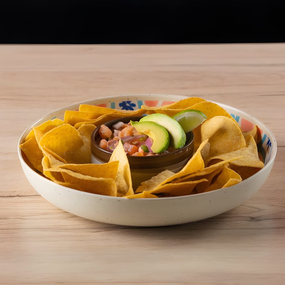 A white bowl filled with tortilla chips surrounds a smaller bowl of pico de gallo, avocado, and lime.